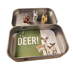 May include: A small, open metal tin with three miniature deer figurines in copper, silver, and gold. The tin's interior features a green card with the text "oh DEER!" and a deer family illustration.