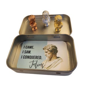 May include: A small, open metal tin containing three miniature busts in copper, silver, and gold. The tin also holds a card with a portrait of Julius Caesar and the quote "I came. I saw. I conquered."