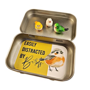 May include: A small tin box with a yellow background and the text "EASILY DISTRACTED BY Birds" inside. The box contains three small bird figurines: a yellow bird, a white bird, and a green bird.