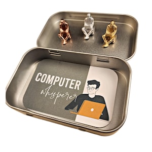 May include: A silver tin with a black and white illustration of a person sitting at a computer with the text "COMPUTER whisperer". The tin contains three small silver, gold, and copper figurines of people sitting at computers.
