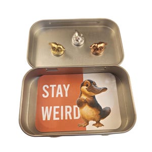 May include: A rectangular metal tin containing three small metallic animal figurines in gold, silver, and copper tones. Inside the base of the tin is a rectangular card with a rust-colored background, the white text STAY WEIRD, and a platypus illustration.