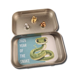 May include: A silver tin with a green snake illustration and the text "2025 Year of the Snake". The tin contains three small silver charms.