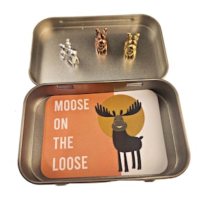 May include: An open metal tin containing three small moose figurines in silver, copper, and gold. A card inside the tin reads "MOOSE ON THE LOOSE" with a cartoon moose illustration on an orange background.
