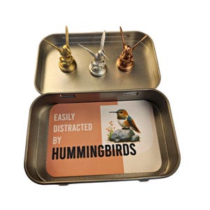May include: An open metal tin with three hummingbird figurines in gold, silver, and copper. Inside, a card reads "EASILY DISTRACTED BY HUMMINGBIRDS" with a hummingbird illustration. The tin is rectangular with rounded corners.