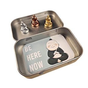 May include: A silver, gold, and copper metal Buddha figurine set in a tin box with a card that reads "Be Here Now".