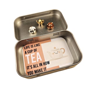 May include: A silver tin with a peach-colored card inside. The card has a black and white illustration of a teapot with a smiling face and the text "Life is like a cup of tea. It's all in how you make it."