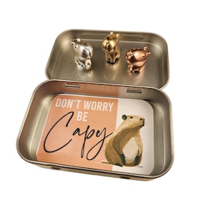 May include: A gold metal tin with a capybara illustration and the text "Don't worry be happy". The tin contains three small capybara figurines, one silver, one gold, and one copper.