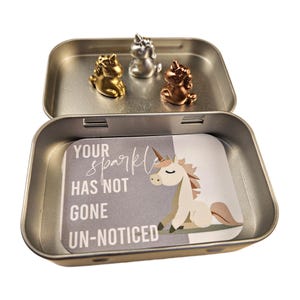 May include: A silver tin with a motivational quote, "Your sparkle has not gone un-noticed." The tin contains three small unicorn figurines, one gold, one silver, and one rose gold.