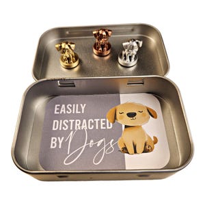 May include: A silver tin with a gray background and the text "Easily Distracted by Dogs". The tin contains three small metal dog figurines in gold, silver, and copper.