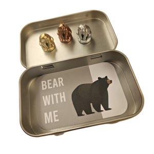 May include: An open silver tin with three small bear figurines in gold, copper, and silver. The tin's interior features a card with a black bear illustration and the text "BEAR WITH ME".