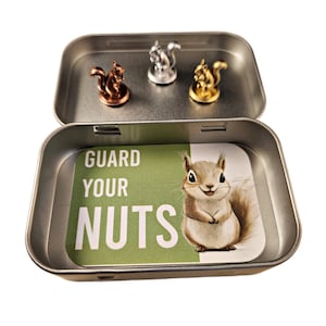 May include: A metal tin with three squirrel figurines in copper, silver, and gold. The tin's interior features a green card with a squirrel illustration and the text "GUARD YOUR NUTS."
