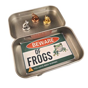 May include: A silver tin with a green and red sign that reads "Beware of Frogs! You have been warned."  Three small frog figurines are visible in the tin, one silver, one gold, and one bronze.