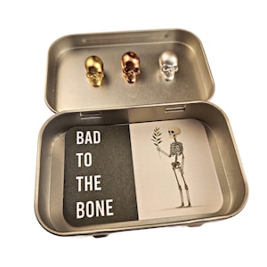 May include: An open silver tin containing three skull-shaped objects in gold, copper, and silver. Inside, two cards display the text "BAD TO THE BONE" and a skeleton illustration holding a sprig.