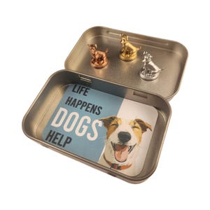 May include: An open metal tin with three small dog figurines in gold, silver, and copper. The tin's interior features a card with a smiling dog and the text "LIFE HAPPENS DOGS HELP".