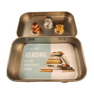 May include: An open, rectangular metal tin with three small, decorative metal objects inside. The tin contains a card with the words "I'll be home reading" and a stack of books. The objects are copper, silver, and gold in color.