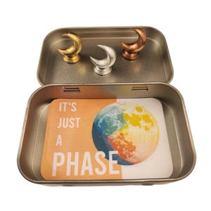 May include: A small, open metal tin containing three crescent moon-shaped objects in gold, silver, and copper. Inside the tin is a card with a colorful illustration of a planet and the text "IT'S JUST A PHASE".