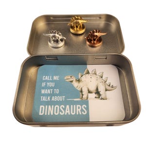 May include: A silver tin containing three dinosaur figurines in gold, silver, and copper. The tin also holds a card with a Stegosaurus illustration and the text "CALL ME IF YOU WANT TO TALK ABOUT DINOSAURS."