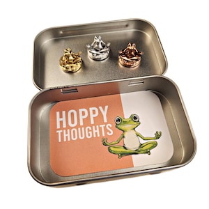 May include: A silver tin with a hinged lid containing three small frog figurines in gold, silver, and copper finishes. The tin has a label that reads "Hoppy Thoughts" and features a green frog in a yoga pose.