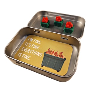 May include: An open, rectangular metal tin containing three miniature green dumpsters with red flames and a yellow card. The card reads "I'M FINE. IT'S FINE. EVERYTHING IS FINE." The tin is on a white background.