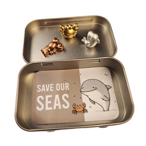 May include: An open metal tin with a printed card and small metal figurines. The card reads "SAVE OUR SEAS" and features a cartoon dolphin and crab. The tin contains a gold crab, silver whale, and bronze lobster.