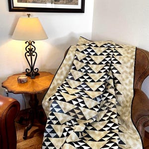May include: A beige and black geometric patterned quilt draped over a brown leather armchair. The quilt features a repeating triangle design. A framed picture of horses hangs on the wall above the chair. A lamp and side table are also visible.
