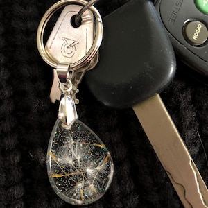 May include: A silver key chain with a teardrop-shaped pendant. The pendant is filled with clear resin and contains a dried dandelion with gold glitter.