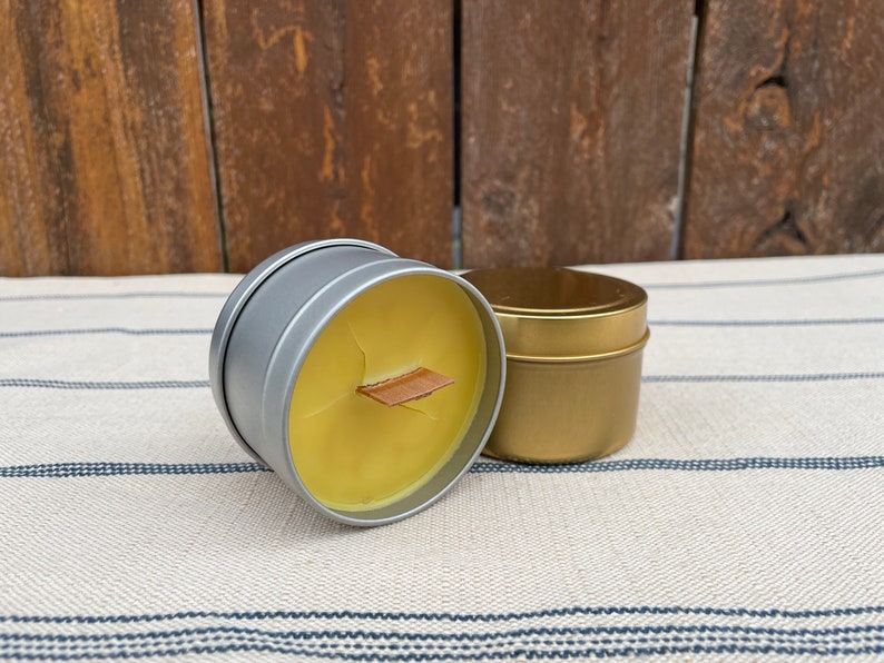 May include: Two small candle tins, one silver and one gold. The silver tin has a wooden wick and is filled with a yellow candle. The gold tin is empty.