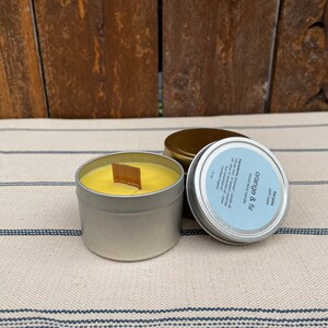May include: A silver tin candle with a yellow wax and a wooden wick. The candle is labeled "Orange & Fir" and "Soy Candle".