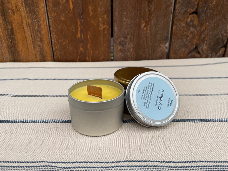 May include: A silver tin candle with a yellow wax and a wooden wick. The candle is labeled "Orange & Fir" and "Soy Candle".
