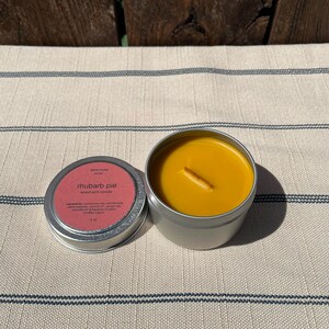 May include: A small, silver tin candle with a yellow wax top and a wooden wick. The label on the tin reads "sara nore studio" and "rhubarb pie" in a pink font. The candle is on a white and black striped surface.
