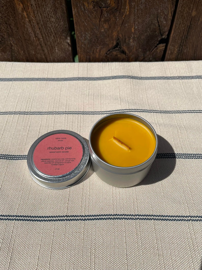 May include: A small, silver tin candle with a yellow wax top and a wooden wick. The label on the tin reads "sara nore studio" and "rhubarb pie" in a pink font. The candle is on a white and black striped surface.