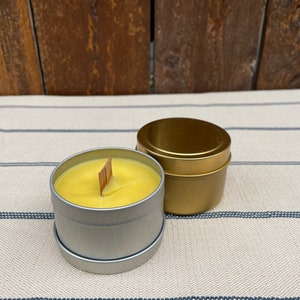 May include: Two candles in metal tins. One candle is yellow and has a wooden wick. The other candle is gold and has a metal lid.
