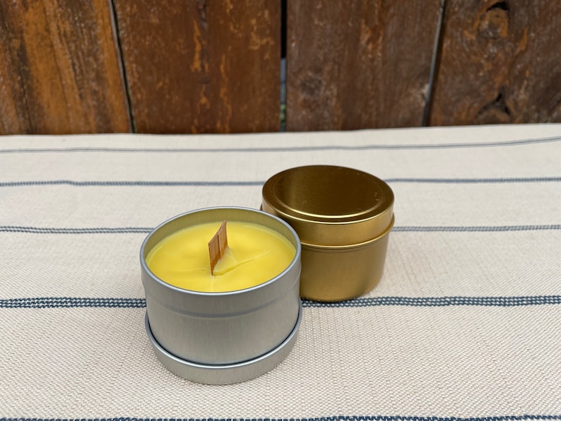 May include: Two candles in metal tins. One candle is yellow and has a wooden wick. The other candle is gold and has a metal lid.