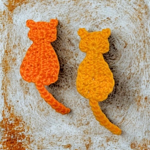 May include: Two crocheted cat shapes in shades of orange and yellow. The cat figures have pointed ears and long tails. The background is a textured, off-white surface.