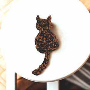May include: A handmade cat-shaped ornament, crafted from brown and black yarn. The cat is depicted from the back, with its tail extended. The ornament is placed on a white surface, showcasing the intricate knit texture.