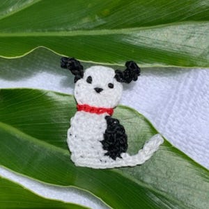 May include: A handmade crochet dog figurine in white and black, with a red collar. The dog has black ears and a black spot on its back. The figurine is set against a green leaf and white background.