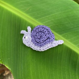 May include: A small, handcrafted crochet snail featuring a light lavender body and a darker purple spiral shell. The snail has two small antennae and a single visible black and white eye, with a textured yarn surface throughout the design.