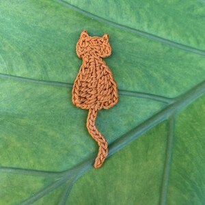 May include: A small, orange, crocheted cat-shaped applique featuring a textured yarn surface, pointed ears, and a long, slender tail extending downward. The piece is crafted with visible interlocking stitches in a solid, warm, earthy orange tone.