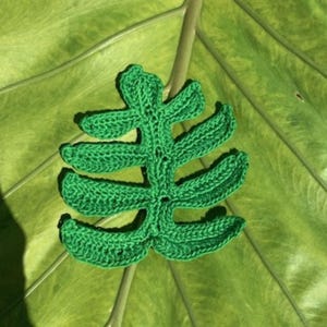 May include: A bright green crochet leaf applique featuring a central stem and four pairs of symmetrical, elongated, pointed lobes extending outward. The textile piece is crafted with a visible yarn stitch pattern, creating a textured, flat, fern-like shape.
