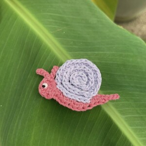 May include: A small, handmade crochet snail figurine featuring a pink body with two antennae and a single black eye. The snail has a prominent, spiraled shell crafted from light lavender yarn, showcasing a textured, circular stitch pattern.