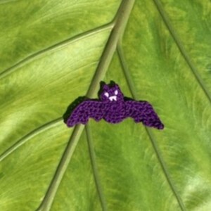 May include: A small, purple crocheted bat applique featuring white yarn details for the eyes and fangs. The bat is depicted with its wings spread wide in a horizontal position, showcasing the textured, knotted yarn construction of the handmade item.