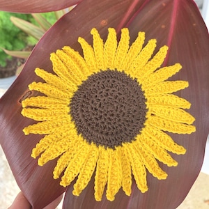 May include: A handcrafted crochet sunflower applique featuring a dark brown circular center surrounded by numerous bright yellow petals. The textile flower is constructed with visible yarn stitches and is displayed against a large, smooth, deep red leaf.