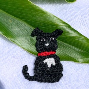 May include: A handmade black crochet dog with a red collar and a white chest marking. The dog is sitting on a white surface with a green leaf in the background. The dog's ears are perked up, and it has a small tail.