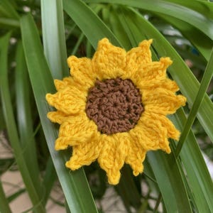 May include: A handcrafted crochet sunflower applique featuring a dark brown circular center surrounded by vibrant yellow petals. The textile flower is constructed with visible yarn stitches, creating a textured, layered appearance with pointed petal tips.