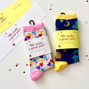 May include: Two pairs of Valentine's Day socks, one with a heart pattern and the other with a space theme. Each pair is wrapped with a band that reads "We make a great pair!" and "Happy Valentine's Day."