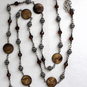 May include: A long silver chain necklace with brown shell and crystal beads. The necklace has a lobster clasp closure.