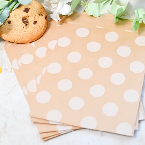 Könnte beinhalten: Ein Stapel brauner Papiertüten mit weißen Punkten. Die Tüten liegen auf einer weißen Oberfläche mit einem Schokoladenkeks und goldenen Konfetti.