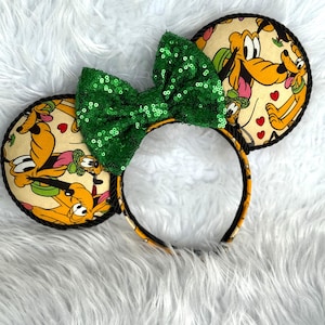 May include: A pair of mouse ears with a green sequined bow. The ears are round with a black rope trim and feature a Pluto cartoon print on a cream background. The headband is black and yellow.