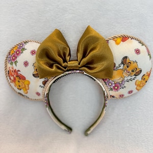 May include: A headband featuring two circular ears with a cream-colored fabric base printed with illustrations of a lion cub, pink flowers, and green leaves. The ears are edged with a twisted tan rope trim and topped with a large, shiny gold bow.