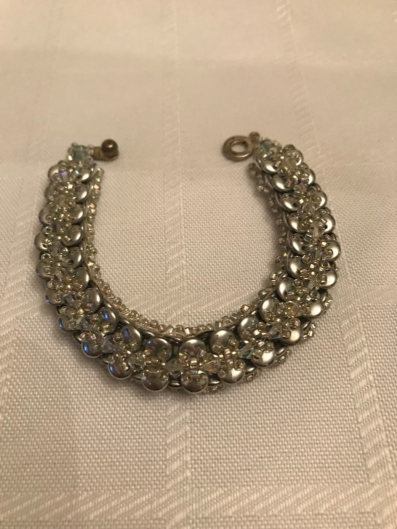 K&ouml;nnte beinhalten: Ein silbernes Armband mit einem Verschluss. Das Armband besteht aus kleinen, runden Perlen, die in einem Muster angeordnet sind. Die Perlen sind silberfarben und haben eine gl&auml;nzende Oberfl&auml;che.